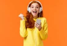 Hooray yes new song. Attractive cheerful and excited redhead woman fist pump in joy and positive emotions, wearing headphones, holding smartphone, smiling camera happily, orange background