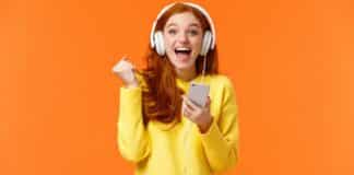 Hooray yes new song. Attractive cheerful and excited redhead woman fist pump in joy and positive emotions, wearing headphones, holding smartphone, smiling camera happily, orange background